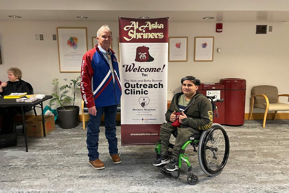 Alejandro, un paciente, en el vestíbulo frente a un cartel que dice "Shriners de Alaska, Bienvenidos a la Clínica de Extensión Walt y Betty Bonner". Hospitales Shriners para Niños - Love to the Rescue Gracias, Centro Médico de Providence, Alaska