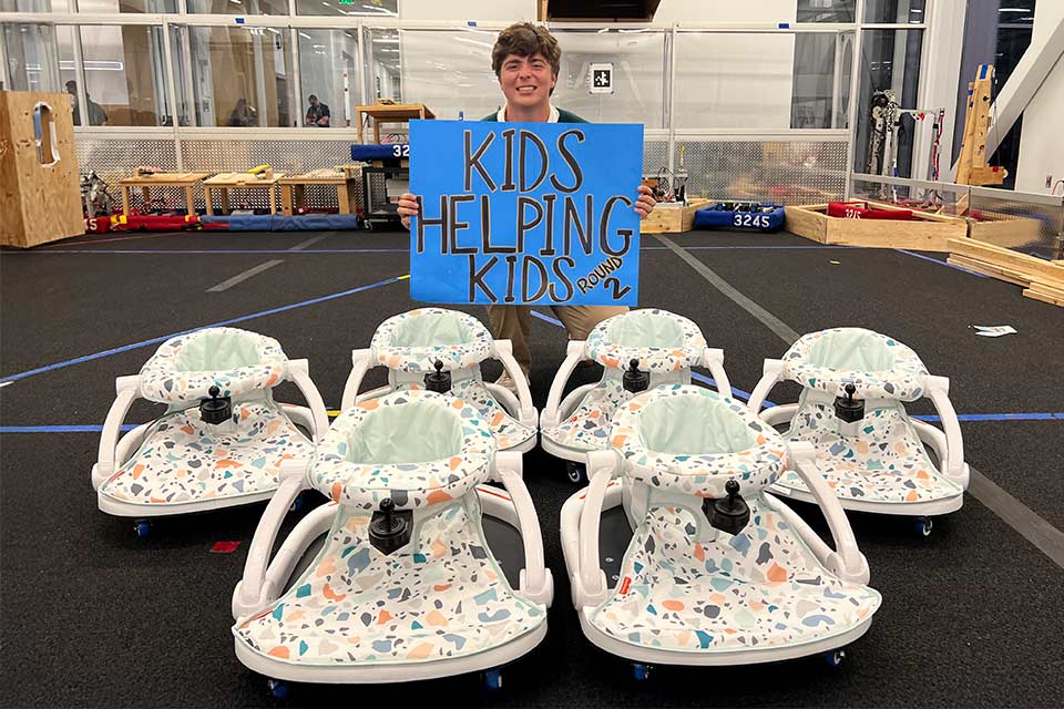 Jack holding a blue sign that reads "Kids Helping Kids Round 2" in front of five Baby Bug Learning Toys.